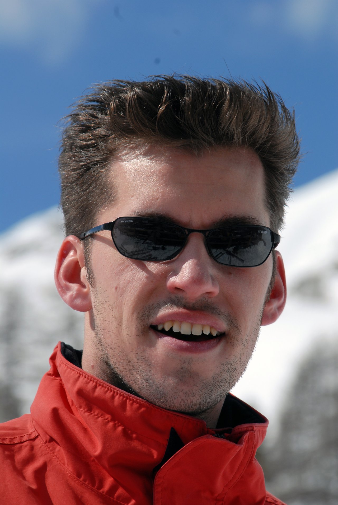 A man wearing sunglasses and a red jacket smiles outdoors with snowy mountains in the background.