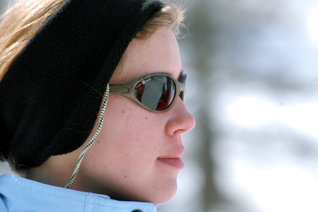 A person wearing sunglasses and a headband looks into the distance, with a snowy background reflected in the lenses.