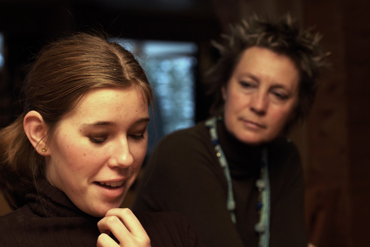 A young woman looks down and smiles while an older woman watches her with a thoughtful expression.