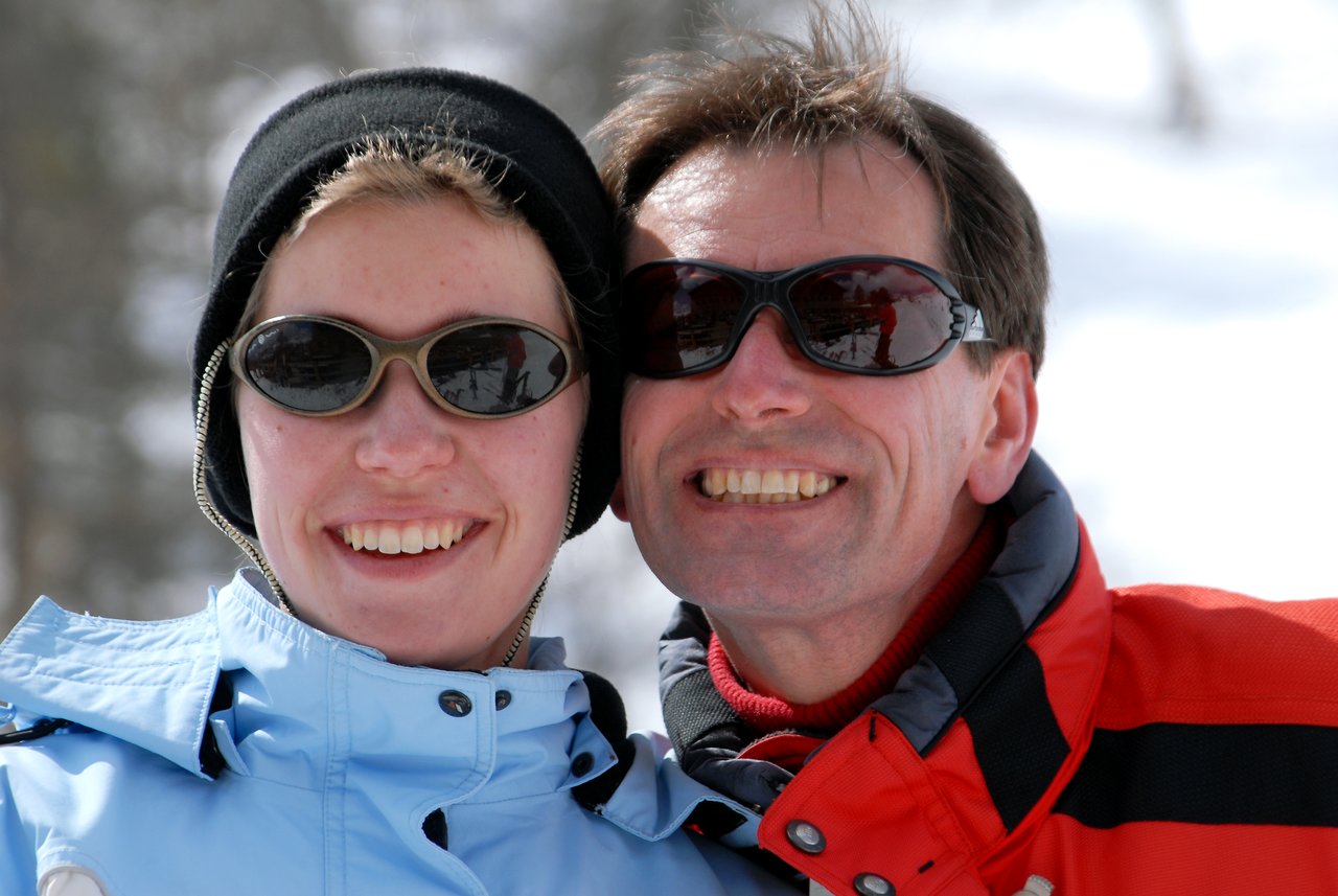 Two people wearing winter jackets and sunglasses smile while posing closely together in a snowy outdoor setting.