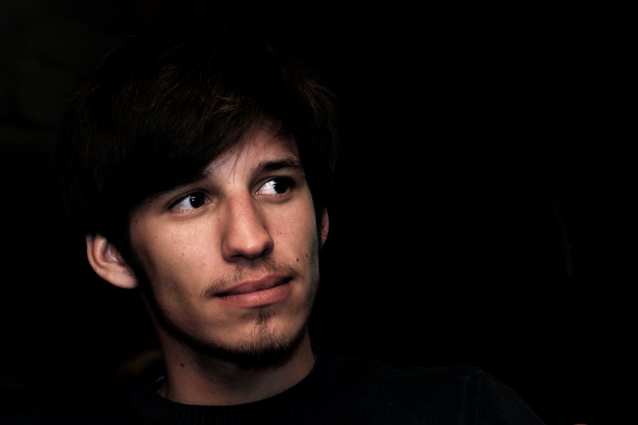 A young man with dark hair and facial hair looks to the side against a dark background.