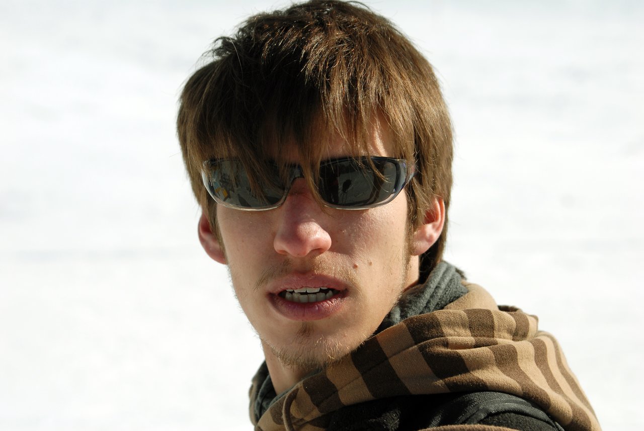 A young man wearing sunglasses and a plaid hoodie looks towards the camera with a neutral expression.