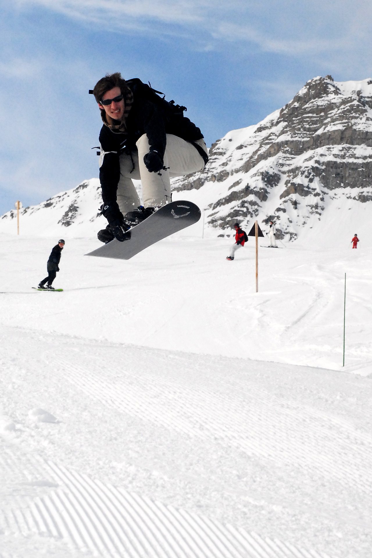 A snowboarder in mid-air performing a jump on a snowy mountain slope, wearing sunglasses and winter gear.