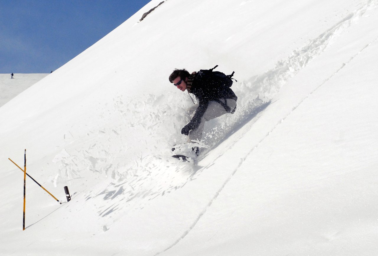 A snowboarder in dark clothing rides down a snowy slope, kicking up powder while making a sharp turn.