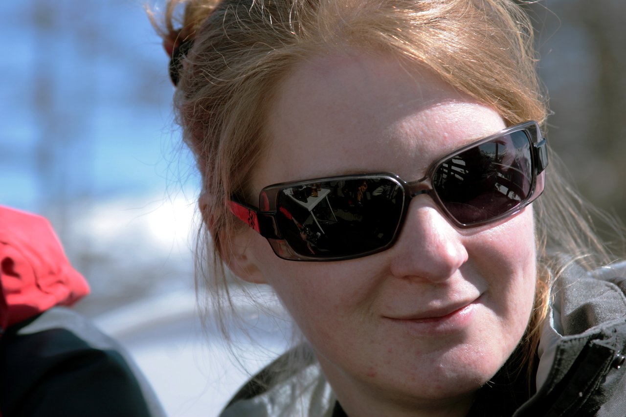 A woman with light skin and sunglasses smiles slightly while outdoors in a snowy setting.