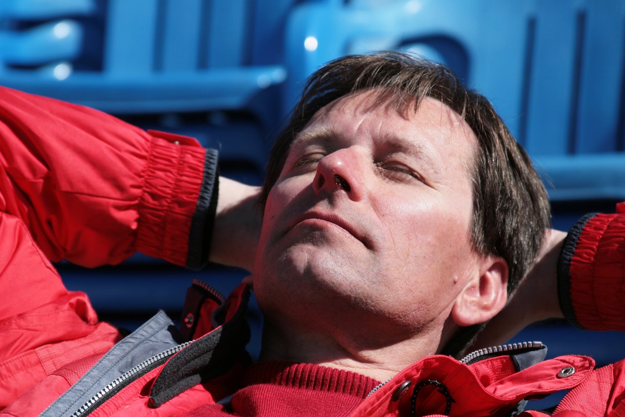 A man in a red jacket reclines with his hands behind his head, eyes closed, enjoying the sunlight.