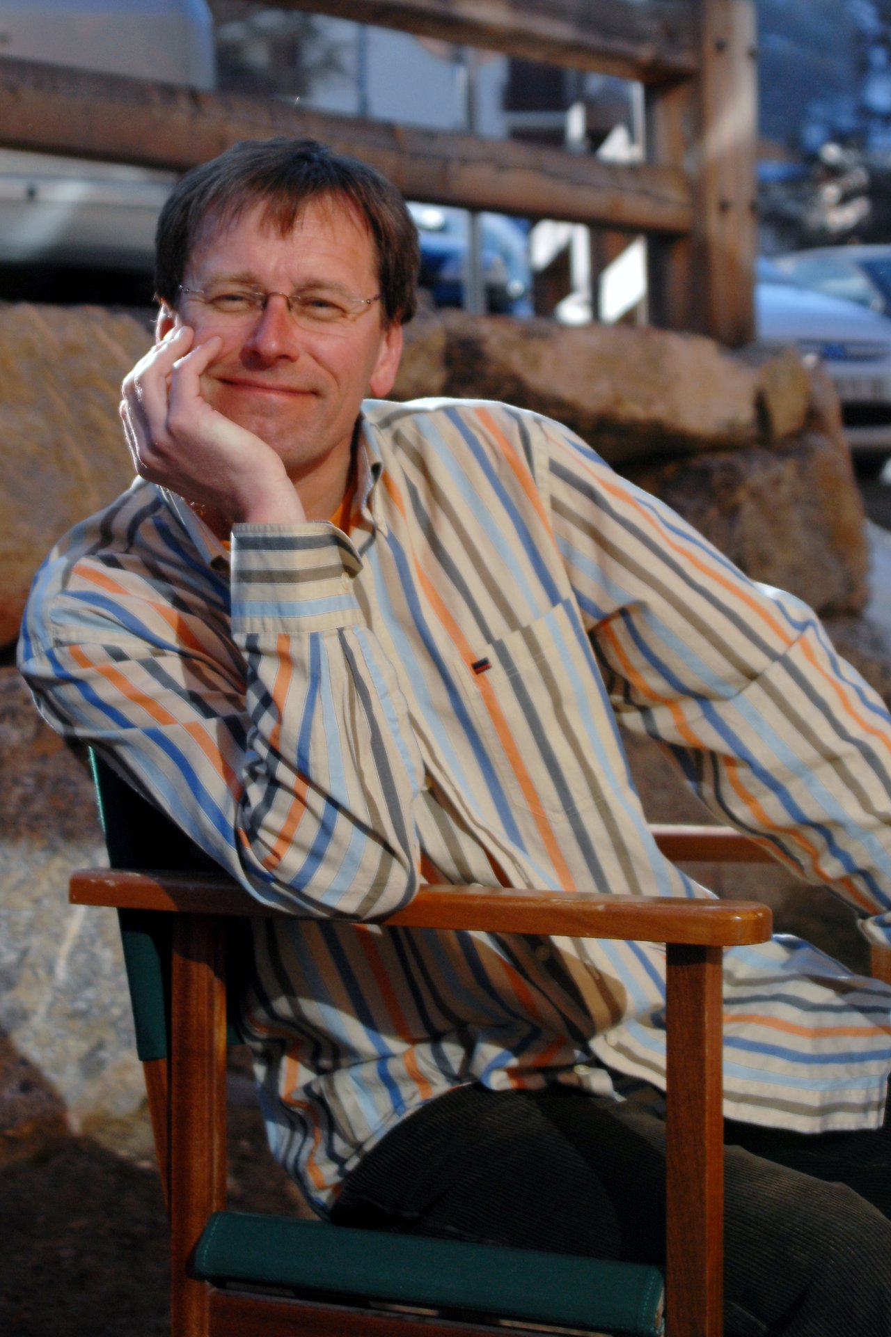 A man wearing glasses and a striped shirt sits on a wooden chair, resting his head on his hand.