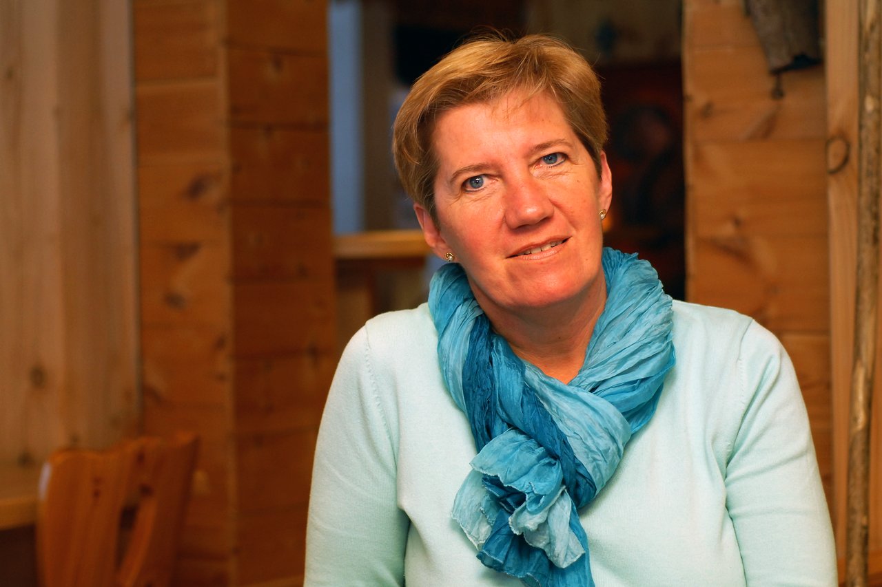 A woman wearing a light blue sweater and blue scarf sits indoors, looking directly at the camera with a slight smile.