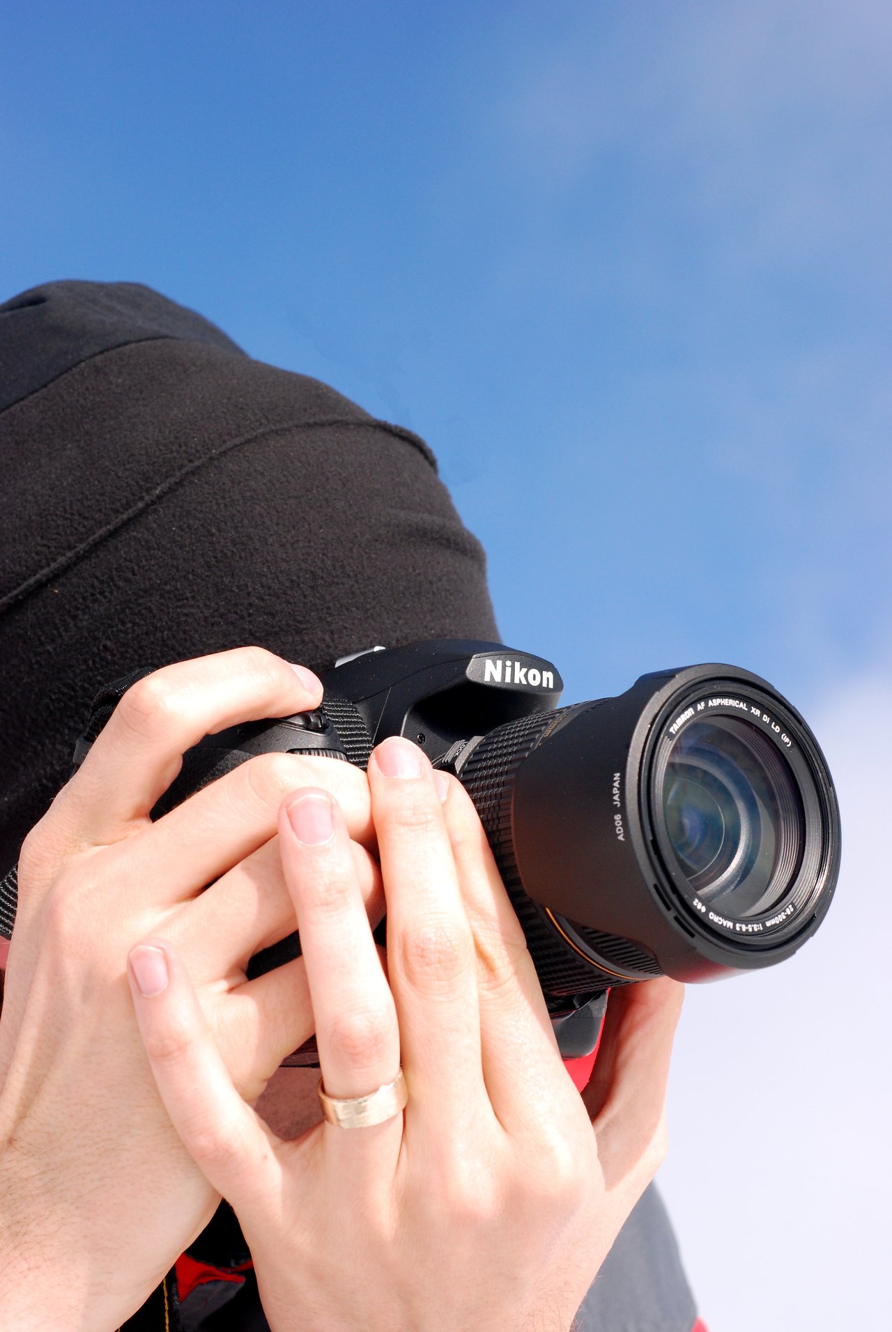 A person wearing a black beanie holds a Nikon camera, focusing on taking a photograph against a clear blue sky.