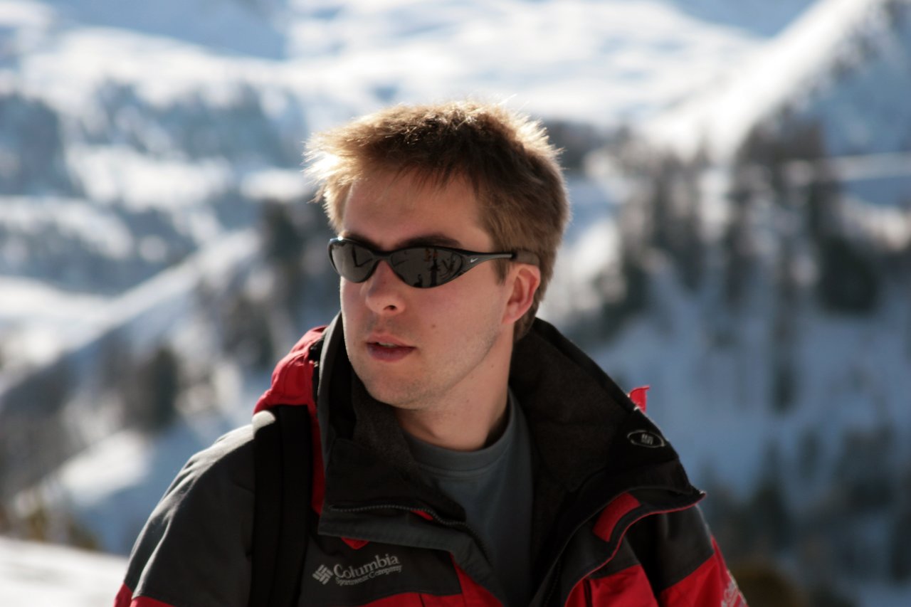 A person wearing sunglasses and a red winter jacket looks to the side with snowy mountains in the background.