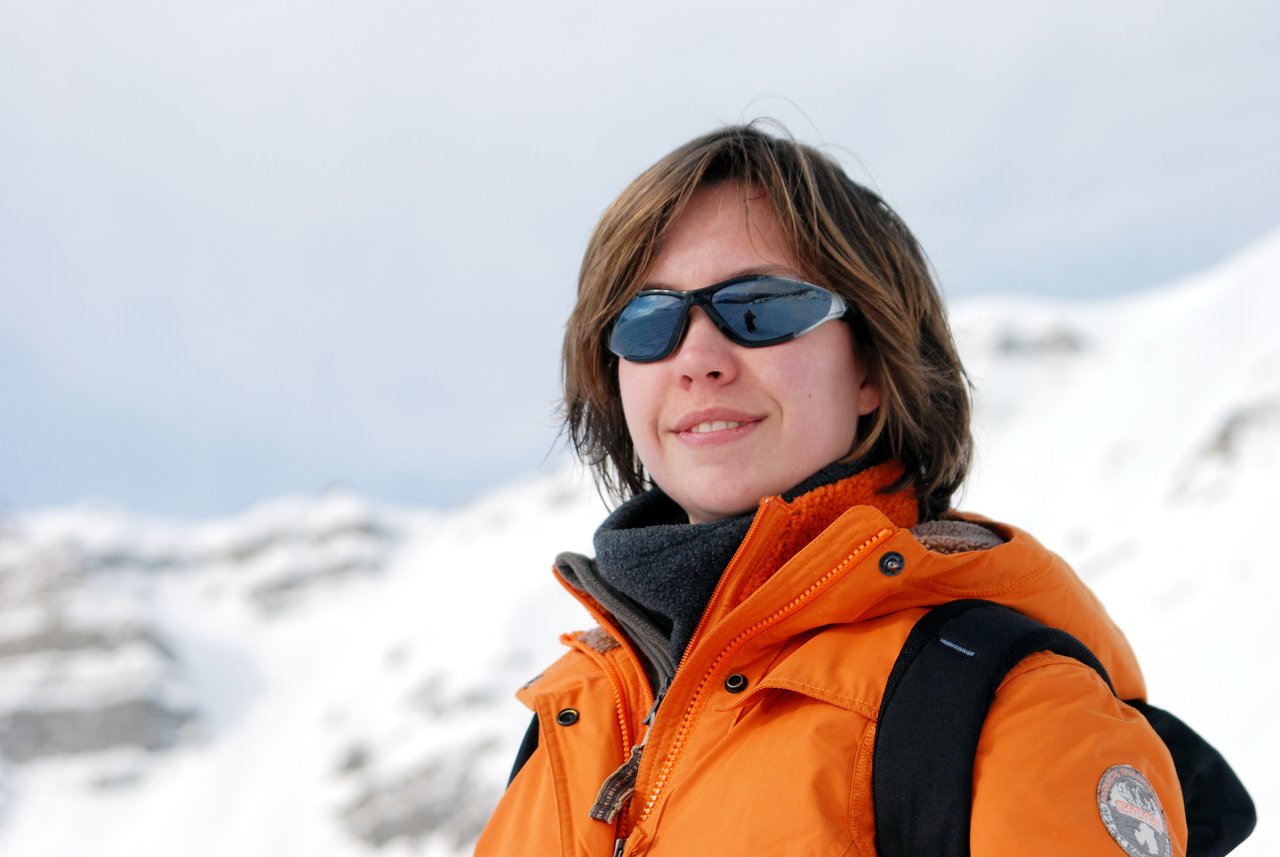 A person wearing sunglasses and an orange jacket stands outdoors in a snowy mountain setting, looking ahead.