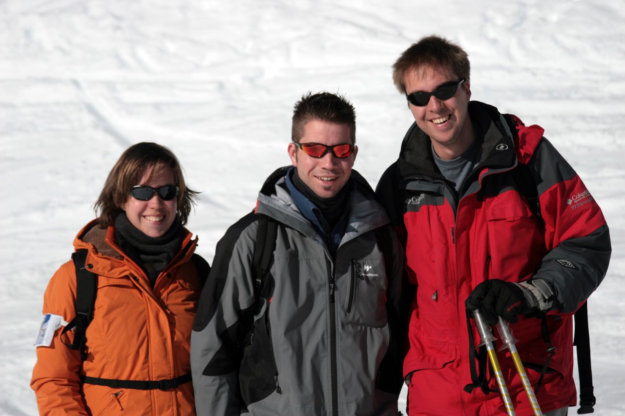 Three people in winter jackets and sunglasses smile while posing on a snowy slope.