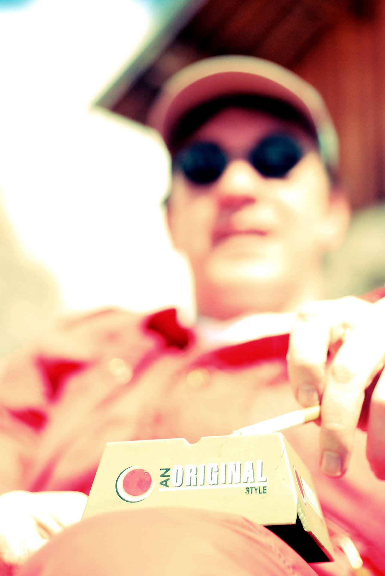 A person in sunglasses and a cap holds a cigarette while taking one from a pack.
