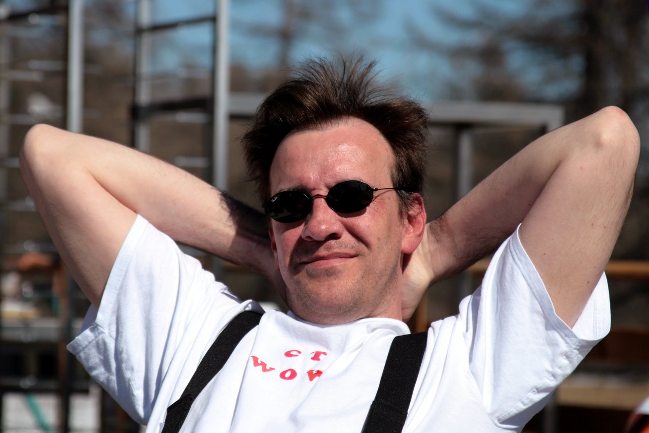 A man wearing sunglasses and a white t-shirt leans back with his hands behind his head, smiling.
