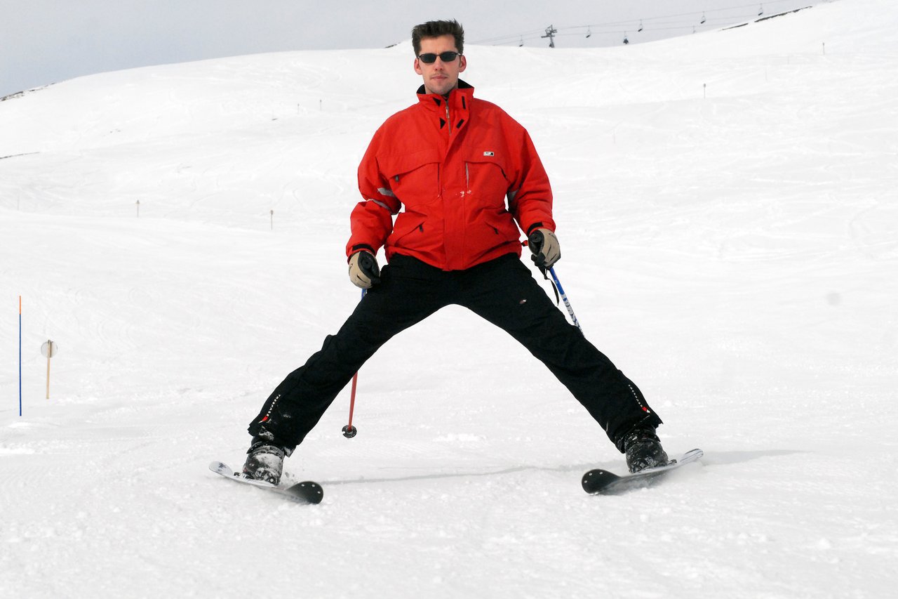 A person in a red jacket and sunglasses skiing with legs wide apart on a snowy slope.
