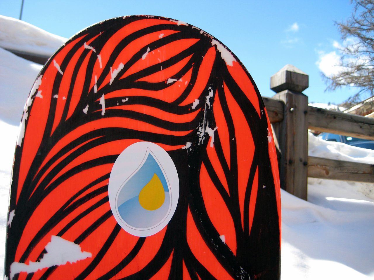 A snowboard with a bold red and black design features an Acquia logo sticker, resting in the snow.