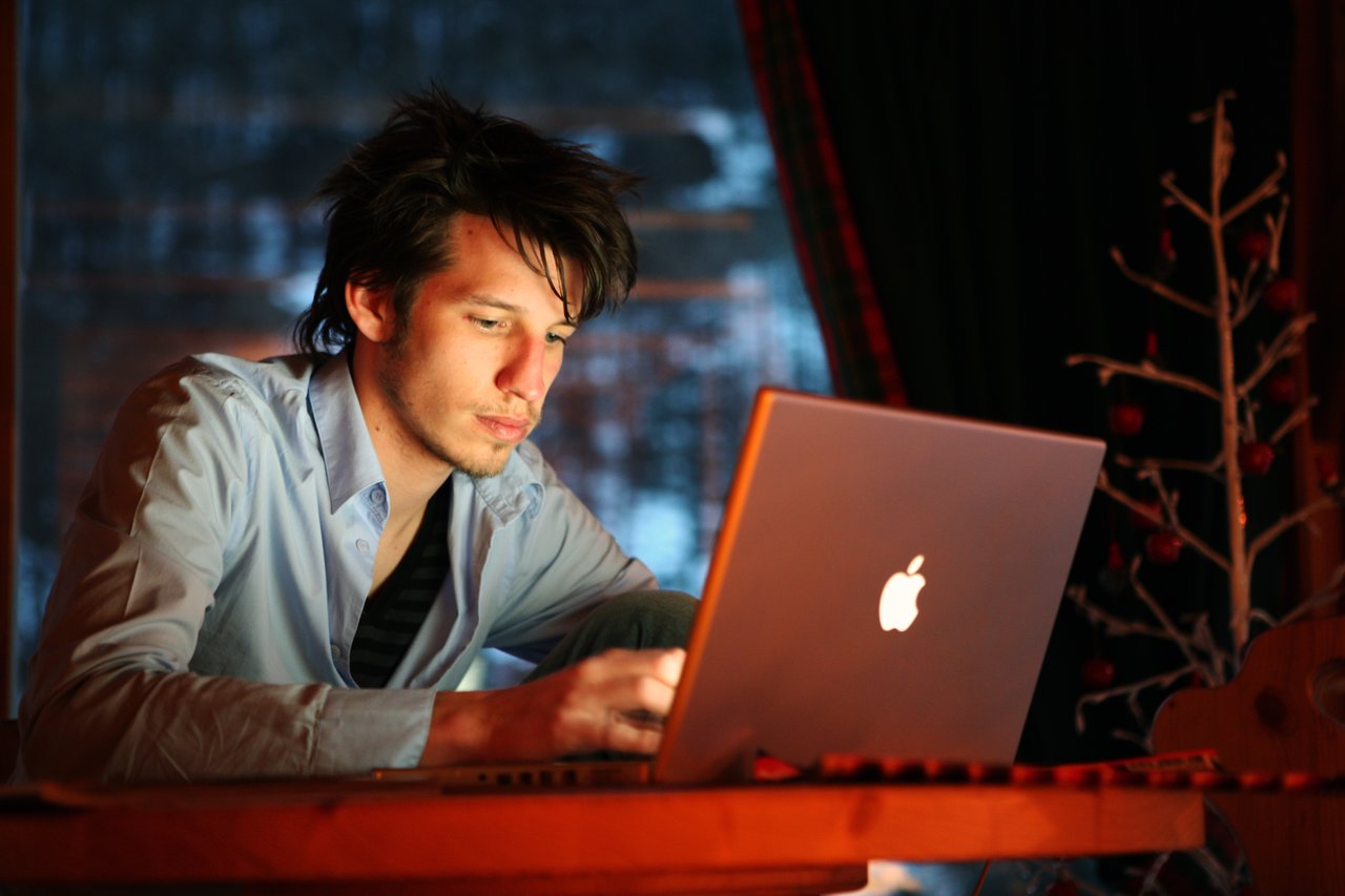 A person in a blue shirt is focused on a laptop, illuminated by its screen in a dimly lit room.