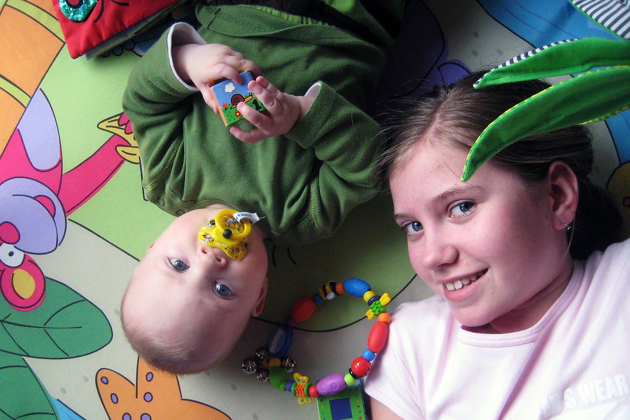 A baby with a pacifier and a young girl lie on a colorful play mat, smiling and playing.