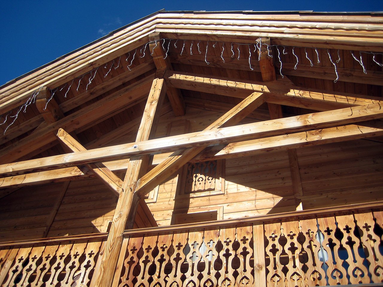 Wooden chalet with decorative balcony railing and exposed beams, viewed from below against a clear blue sky.