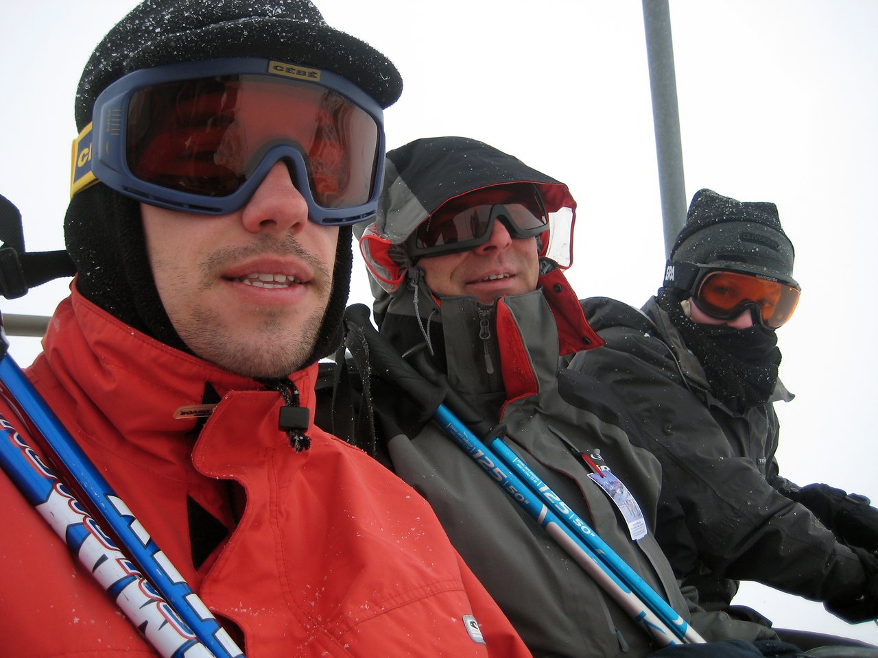 Three people in ski gear sit on a ski lift, wearing goggles and winter jackets in snowy conditions.