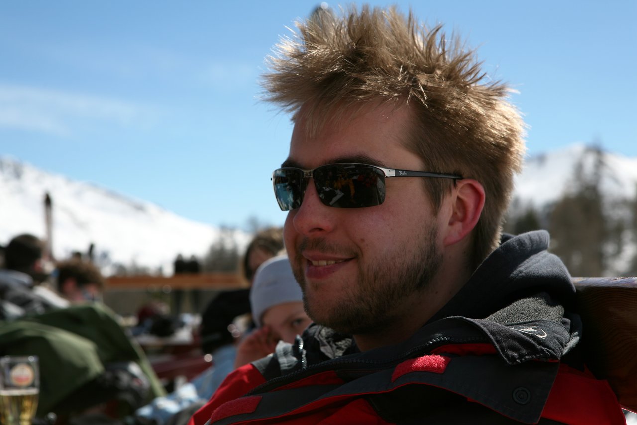 A man wearing sunglasses and a winter jacket smiles while sitting outdoors with snowy mountains in the background.