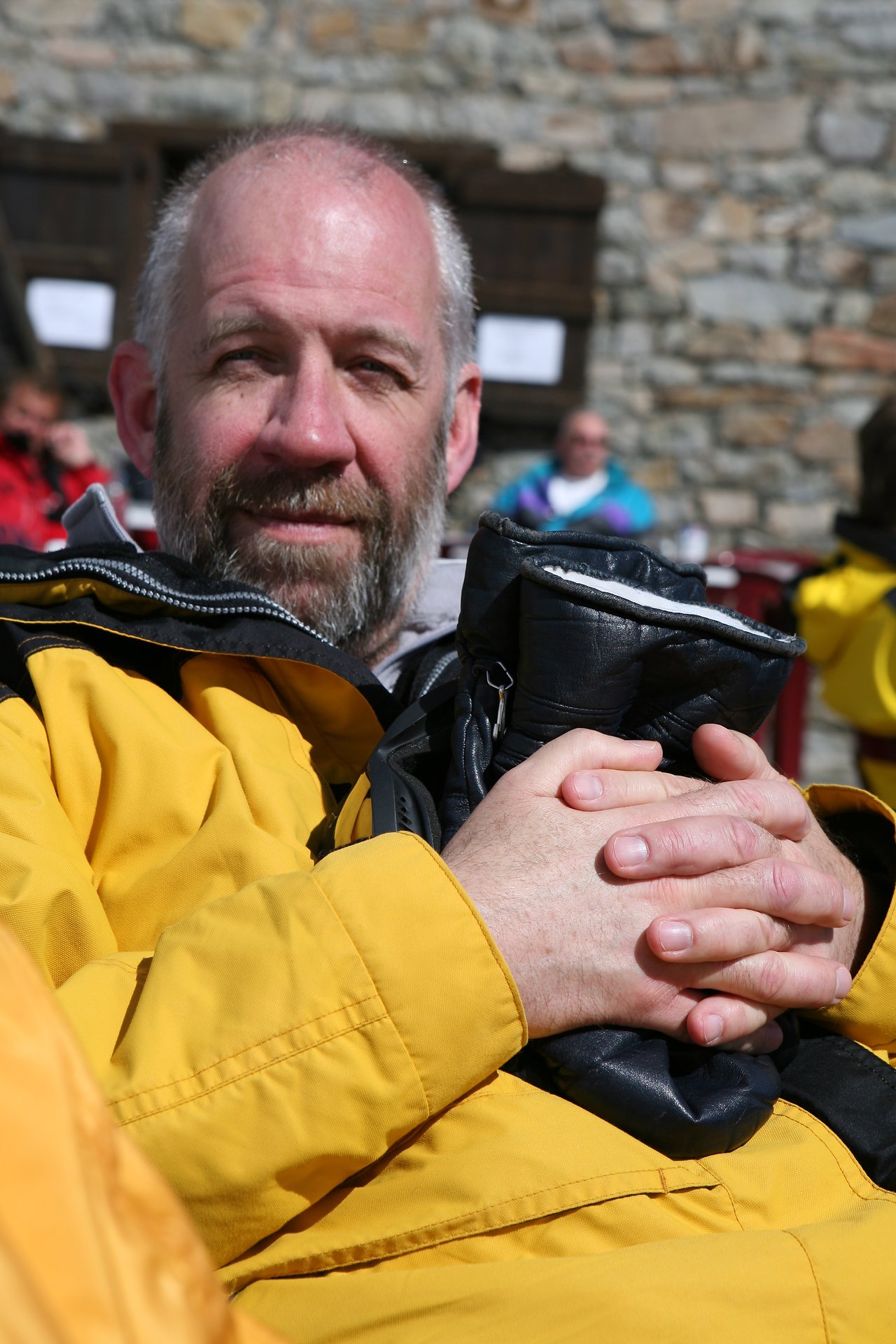 A man in a yellow jacket sits outdoors, holding black gloves in his hands and looking at the camera.