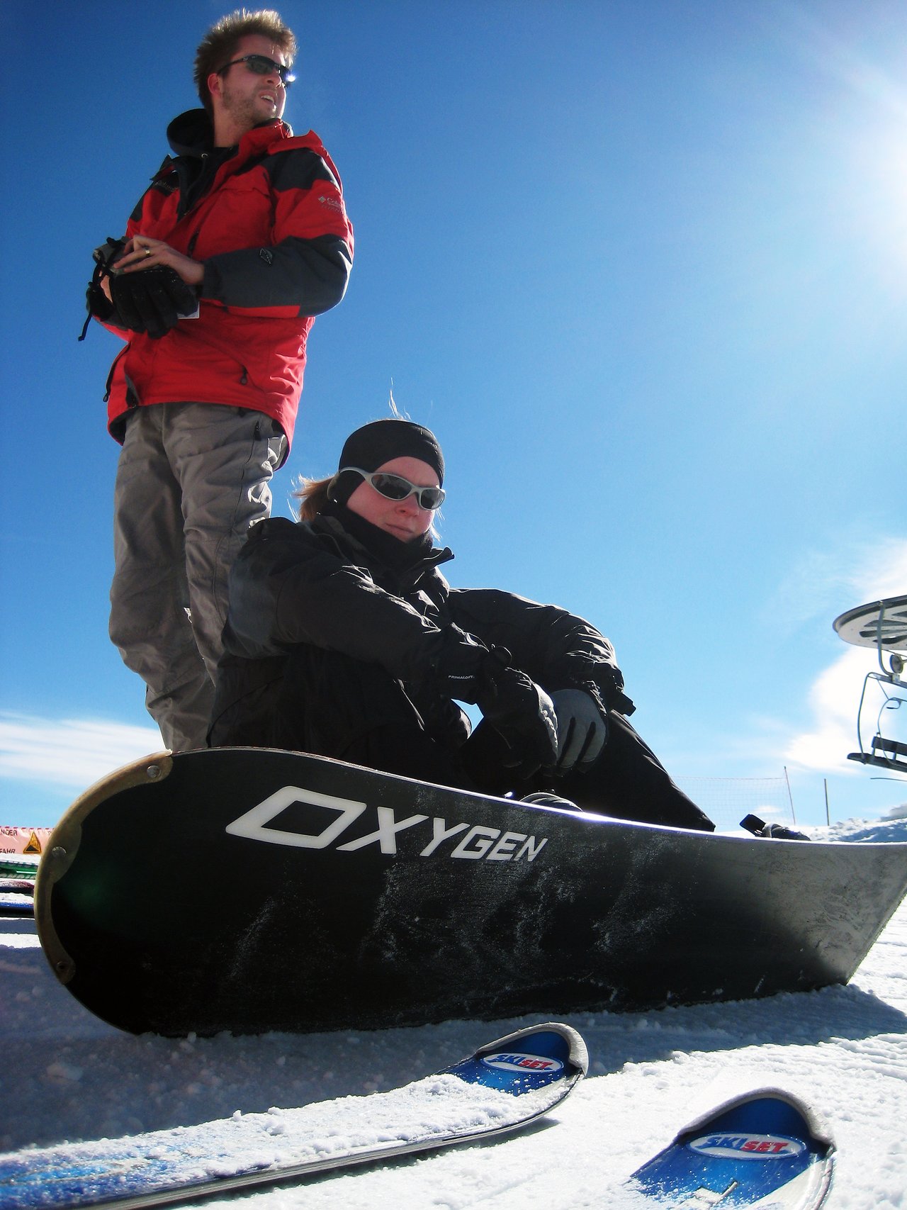 Two people in winter gear on a snowy slope, one sitting with a snowboard and the other standing nearby.