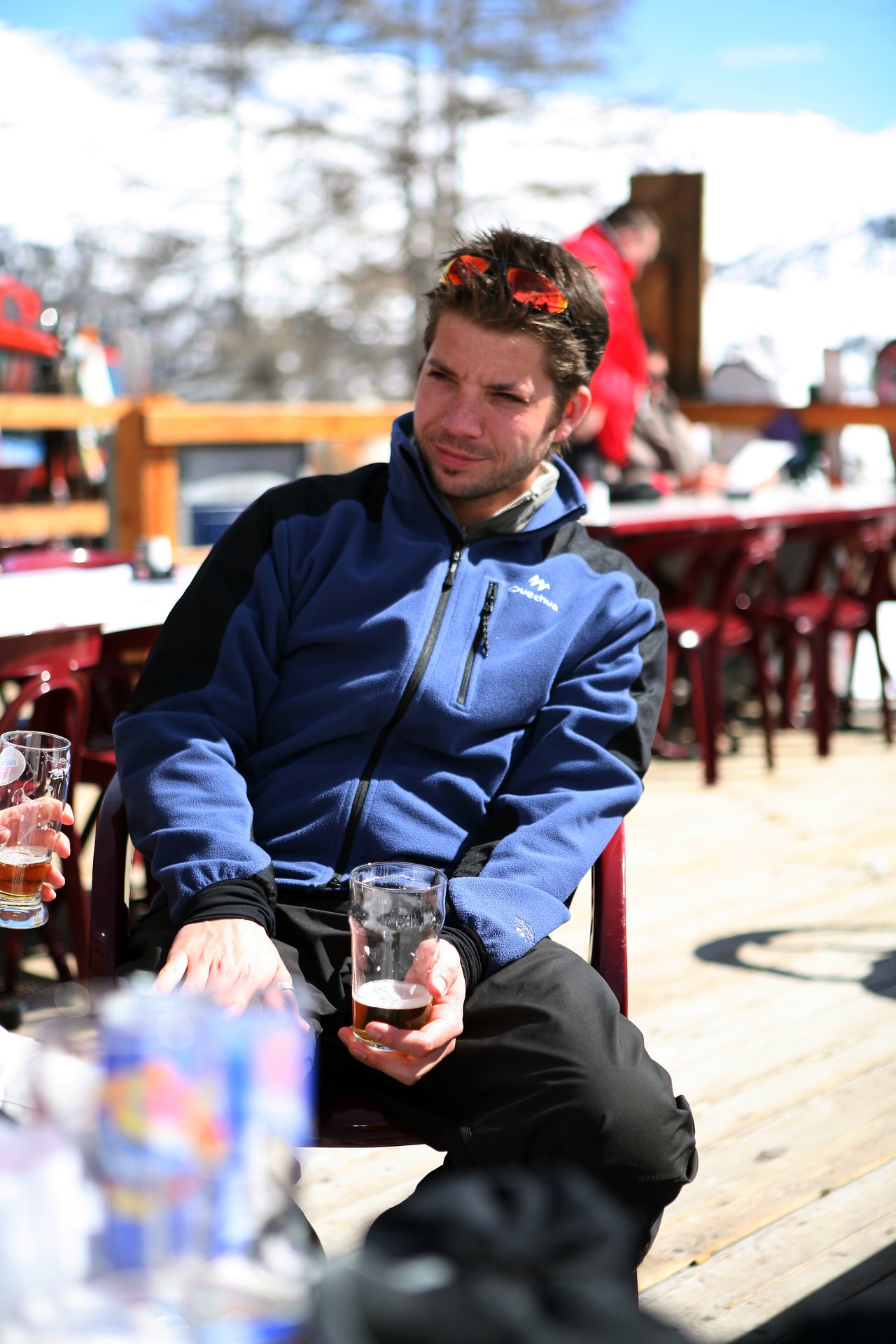 A man in a blue jacket sits outdoors, holding a glass with beer, squinting in the sunlight.