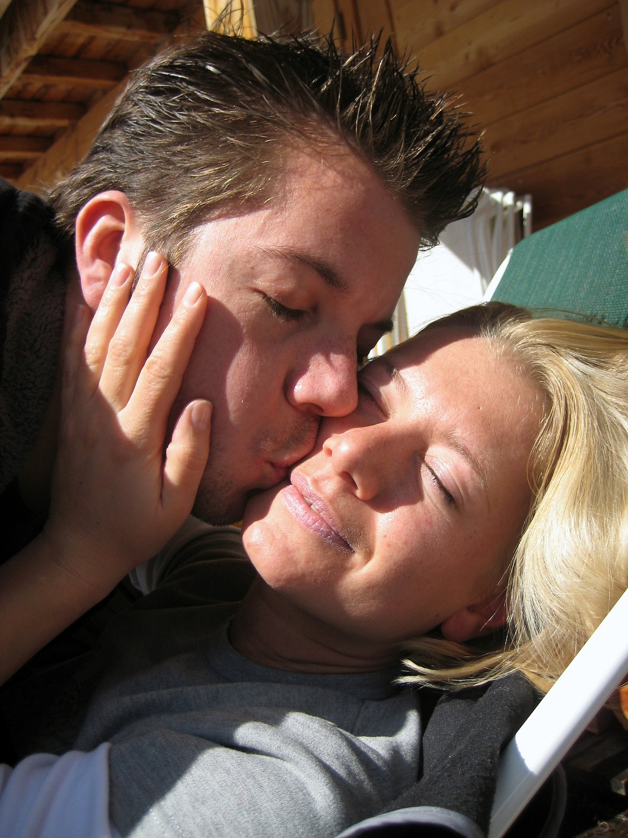 A person kisses another on the cheek while they smile and relax in the sunlight.