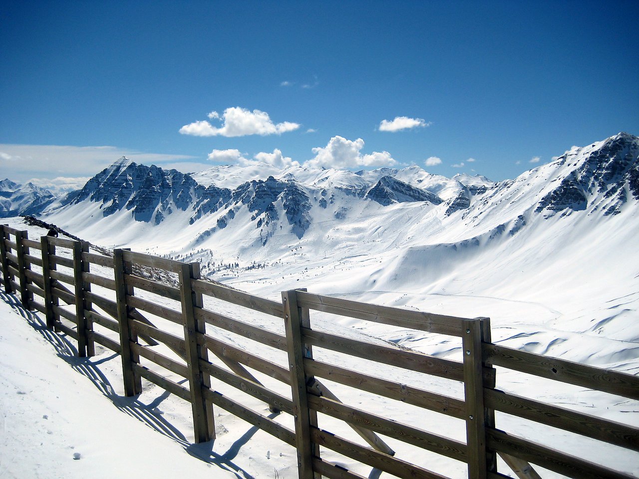 A wooden fence runs along a snowy mountain slope with snow-covered peaks in the background under a clear blue sky.