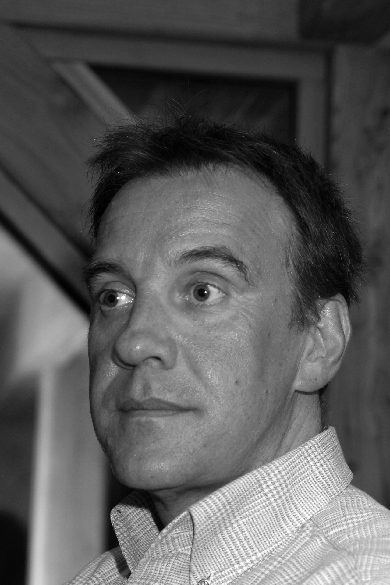 A man with short hair and a checkered shirt looks to the side with a neutral expression.