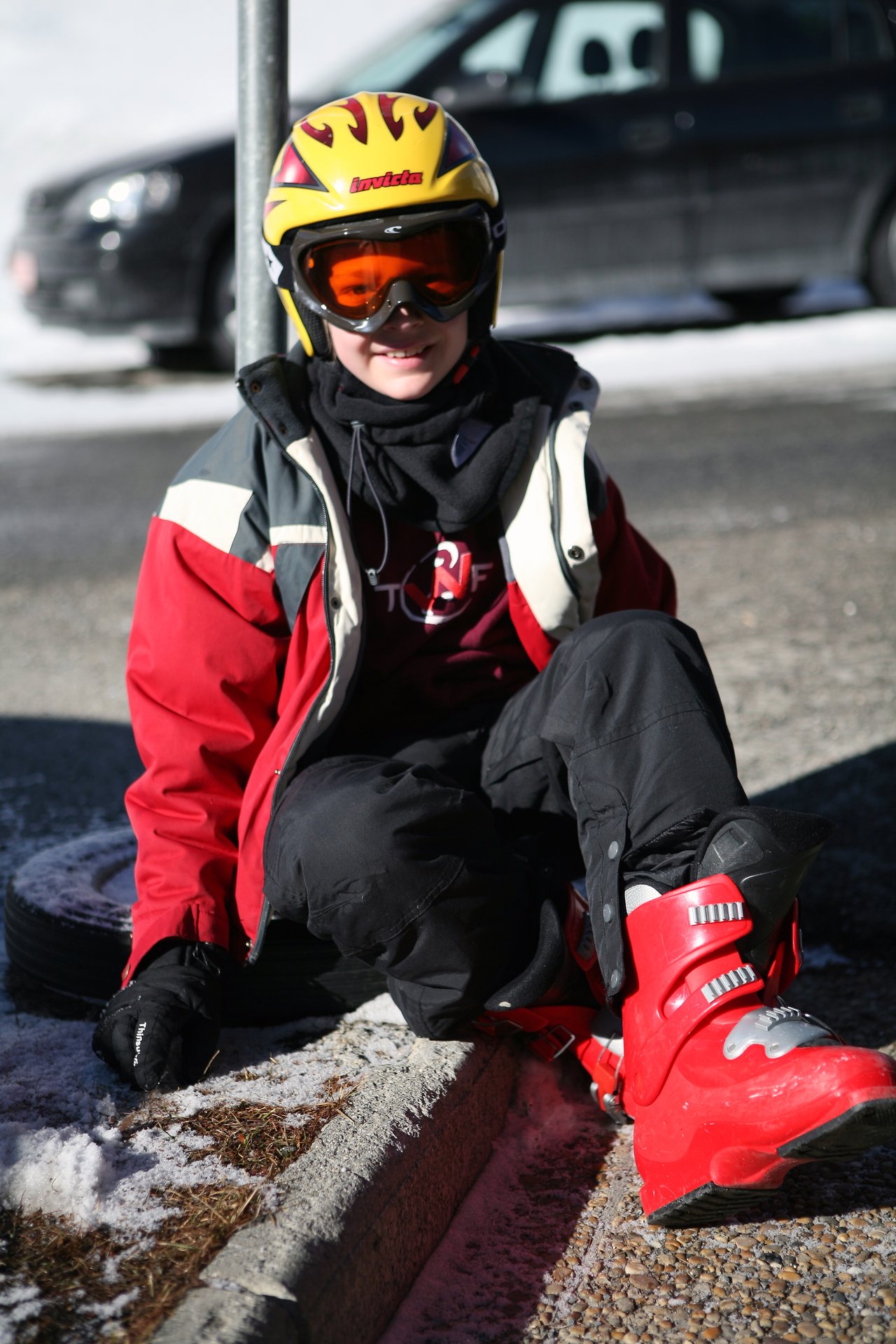 A person in ski gear sits on the ground, wearing a helmet, goggles, and red ski boots.
