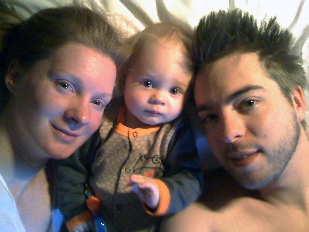 A couple and their baby lie in bed together, looking at the camera.