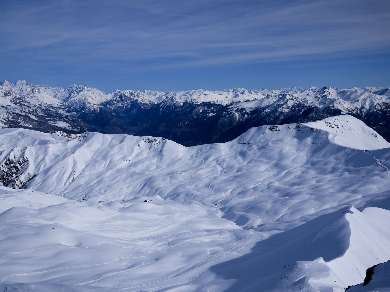 Snow-covered mountain slopes with ski tracks, surrounded by distant peaks under a clear blue sky.