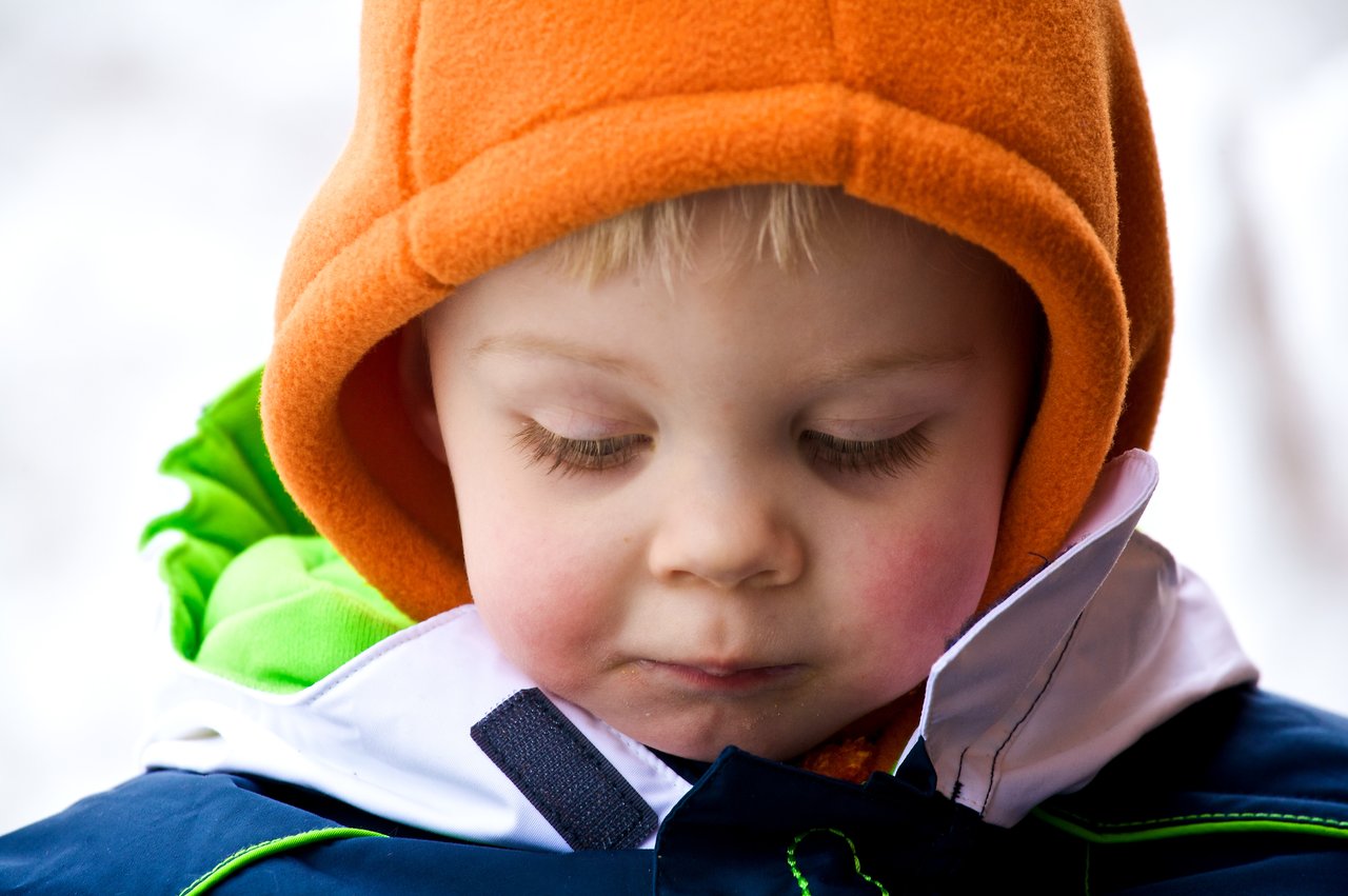 A young child in an orange hood looks down with a calm expression, wearing a winter jacket.