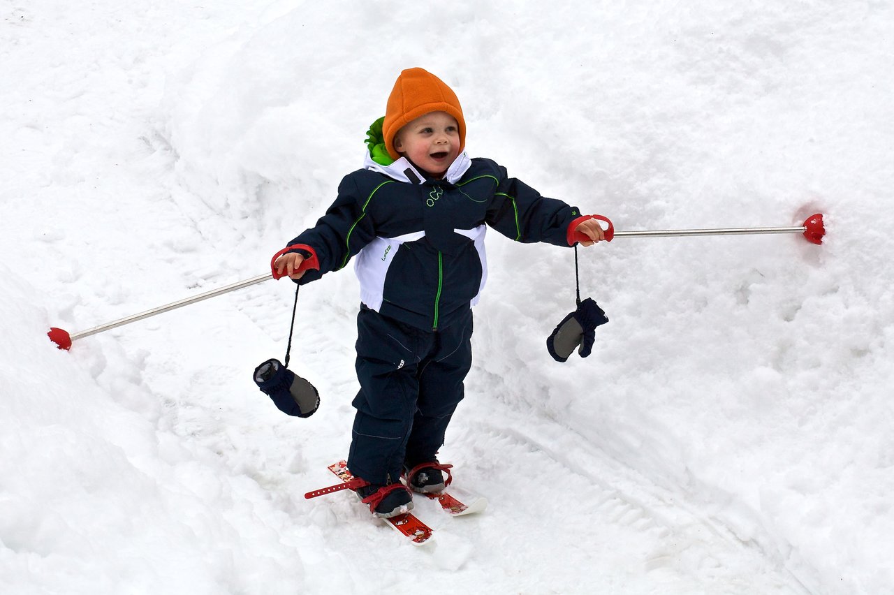 A young child in winter gear skis on snow, holding ski poles with mittens attached.