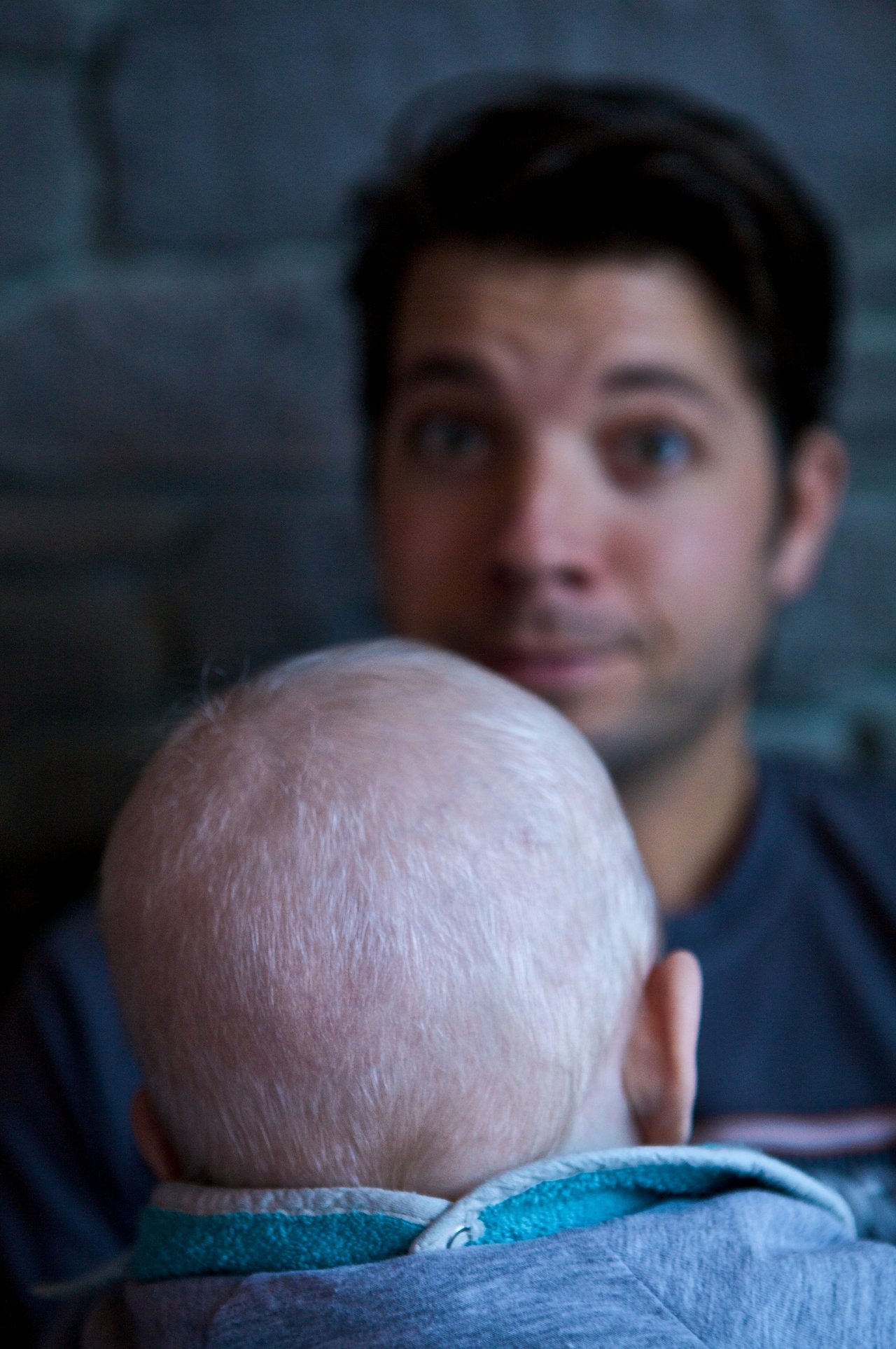 A baby with a light-colored head is seen from behind, facing an adult with a surprised expression.