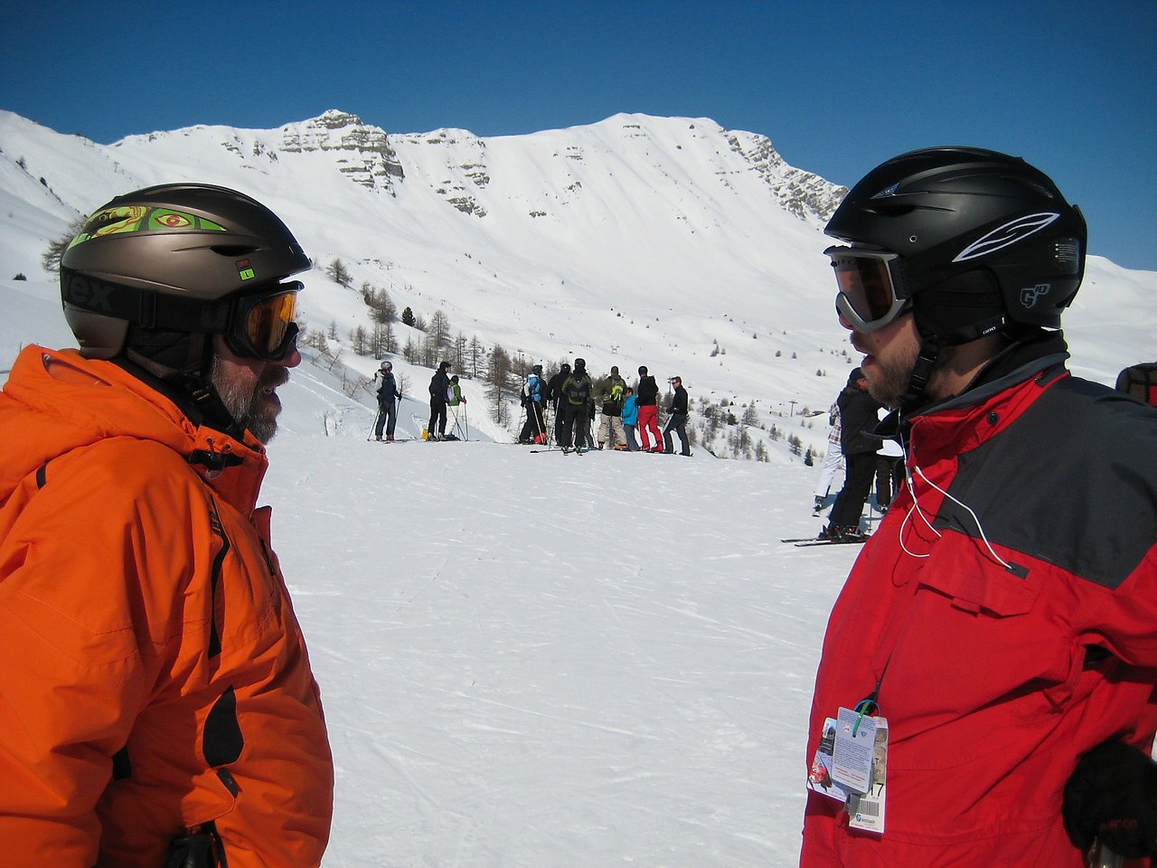 Two people in ski gear stand facing each other on a snowy mountain, with other skiers in the background.