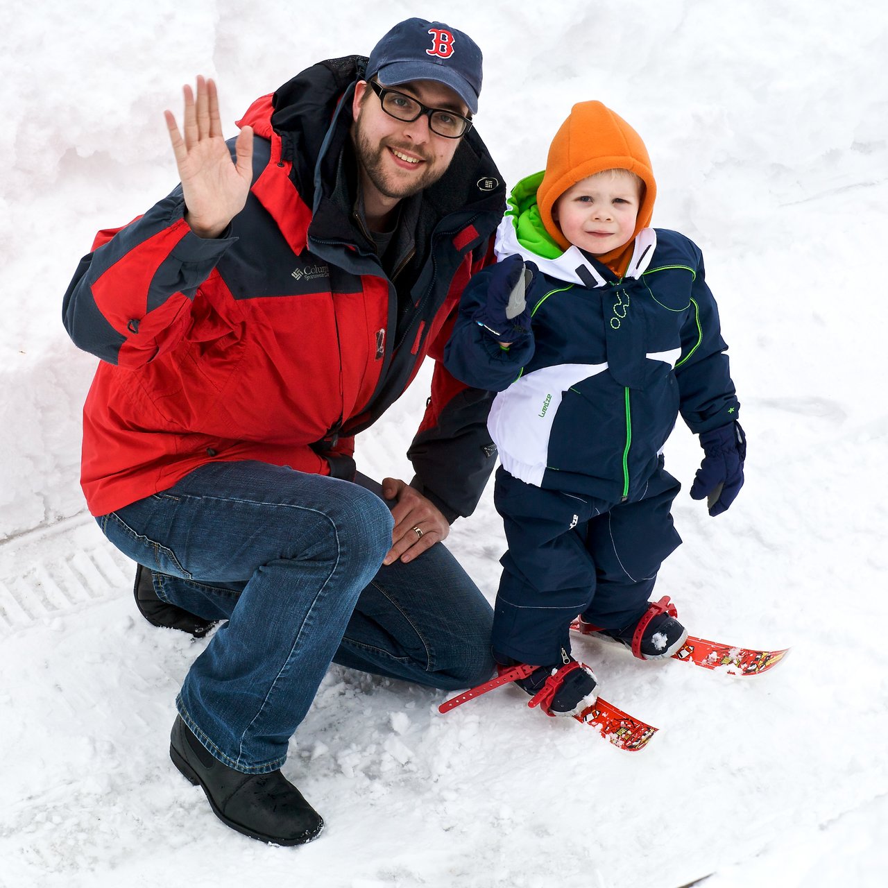 A man and a young child in winter clothing wave while kneeling and standing on snow, with the child wearing skis.
