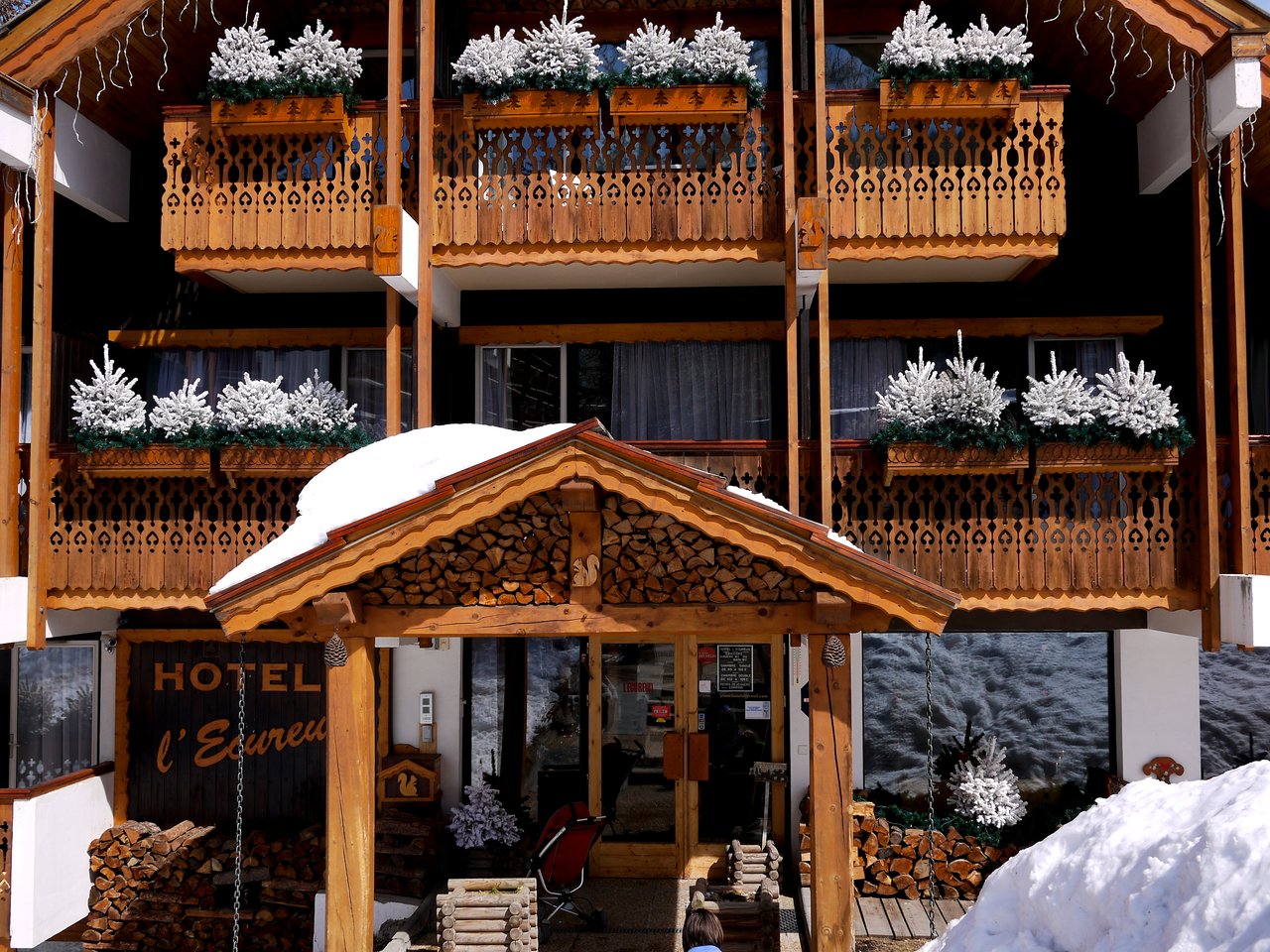 A wooden alpine hotel with snow-covered balconies, decorative plants, and a log-covered entrance under a snowy roof.