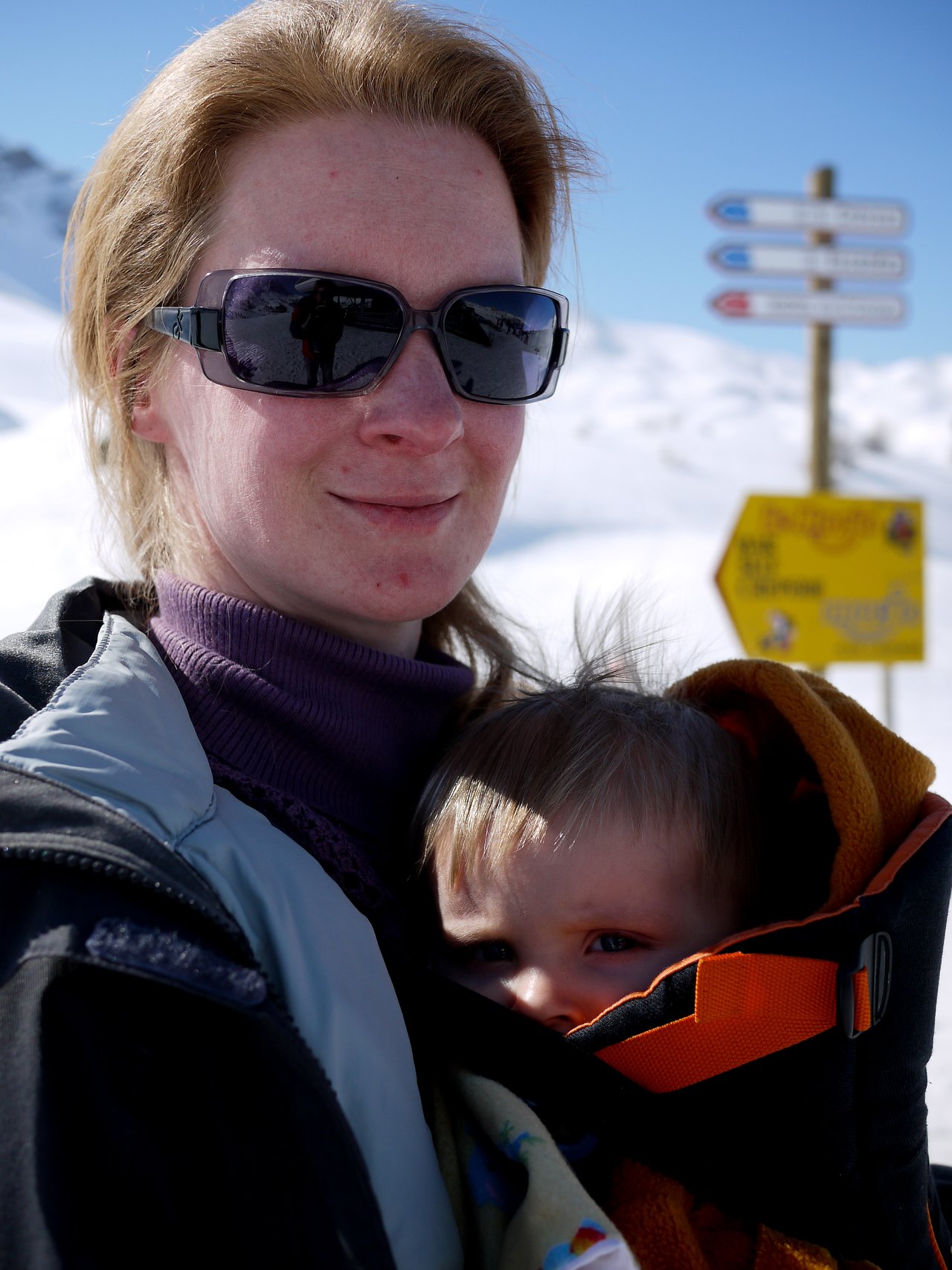 A woman wearing sunglasses carries a baby in a front carrier while outdoors in a snowy environment.