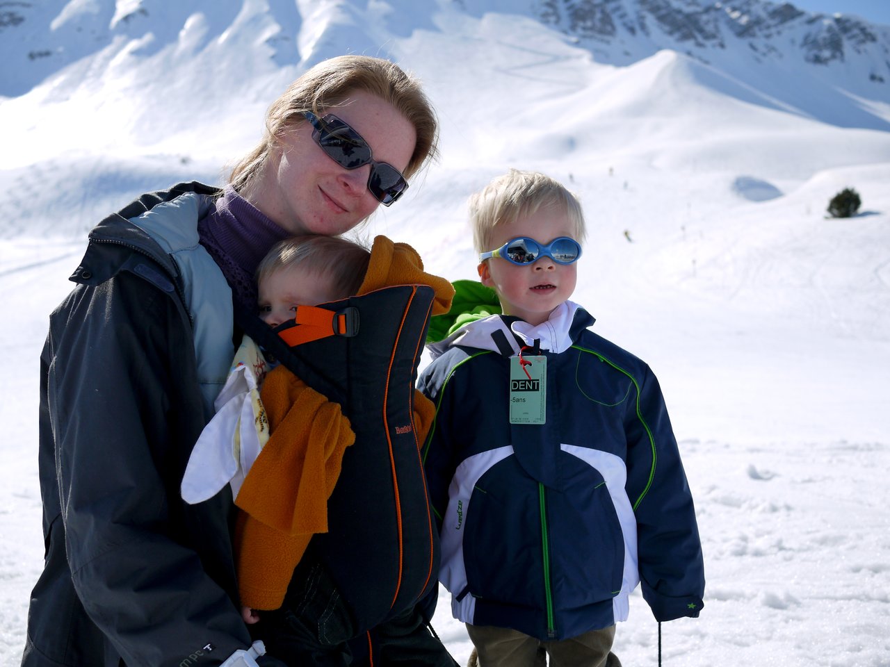 A woman wearing sunglasses holds a baby in a carrier, while a young child in winter gear stands nearby.
