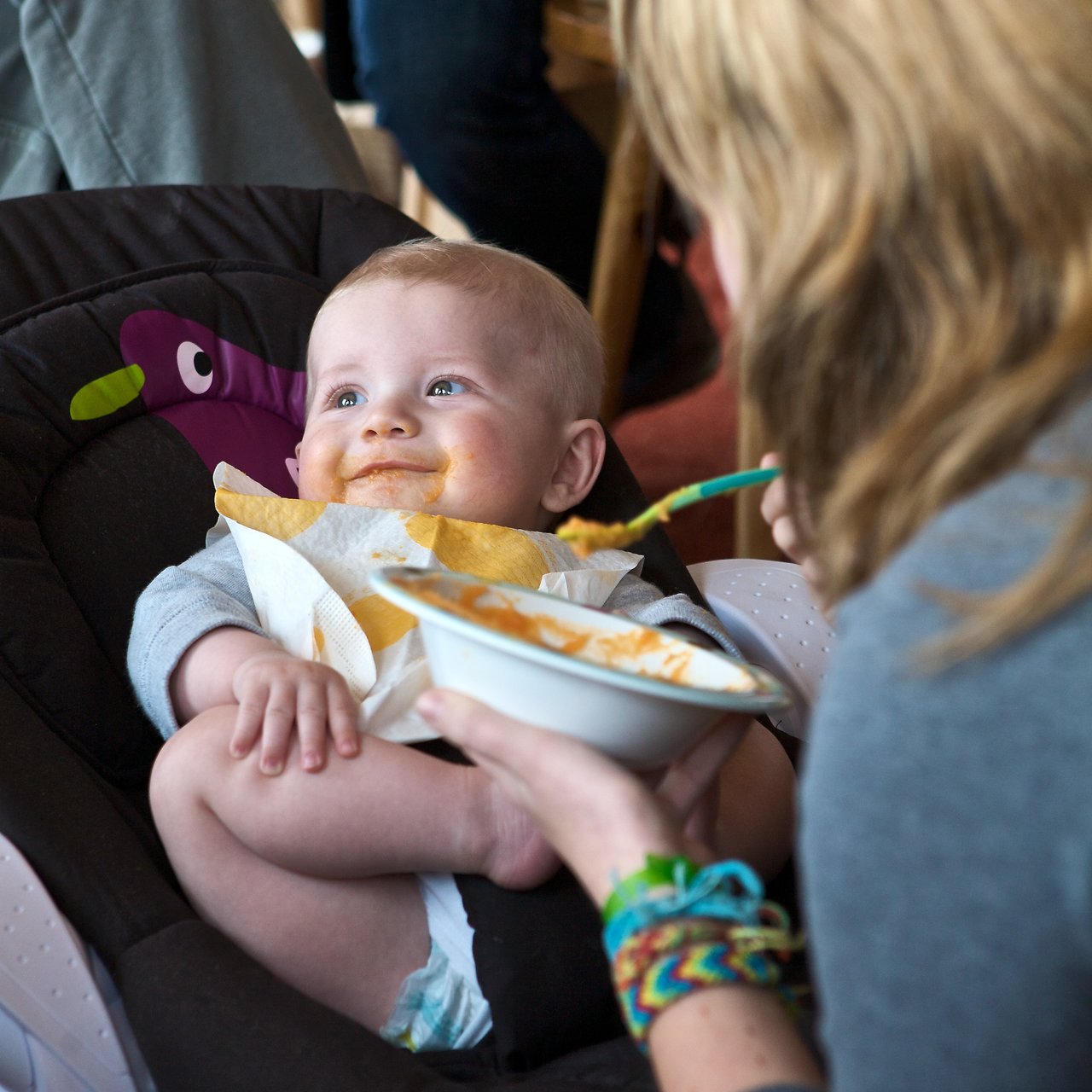 A baby with food on their face sits in a high chair, smiling while being fed by an adult.