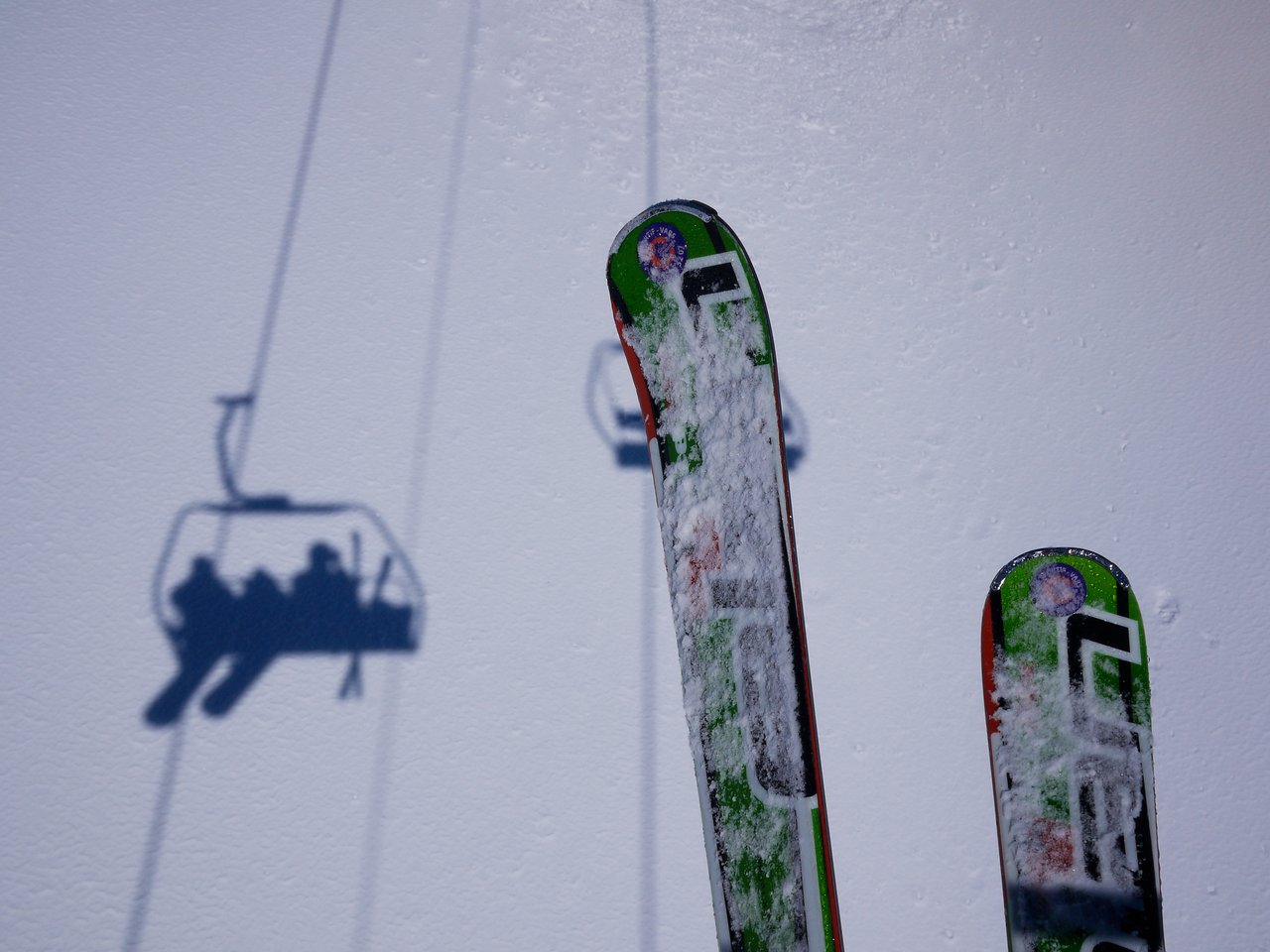 A pair of snow-covered skis with a ski lift shadow on the snowy ground.