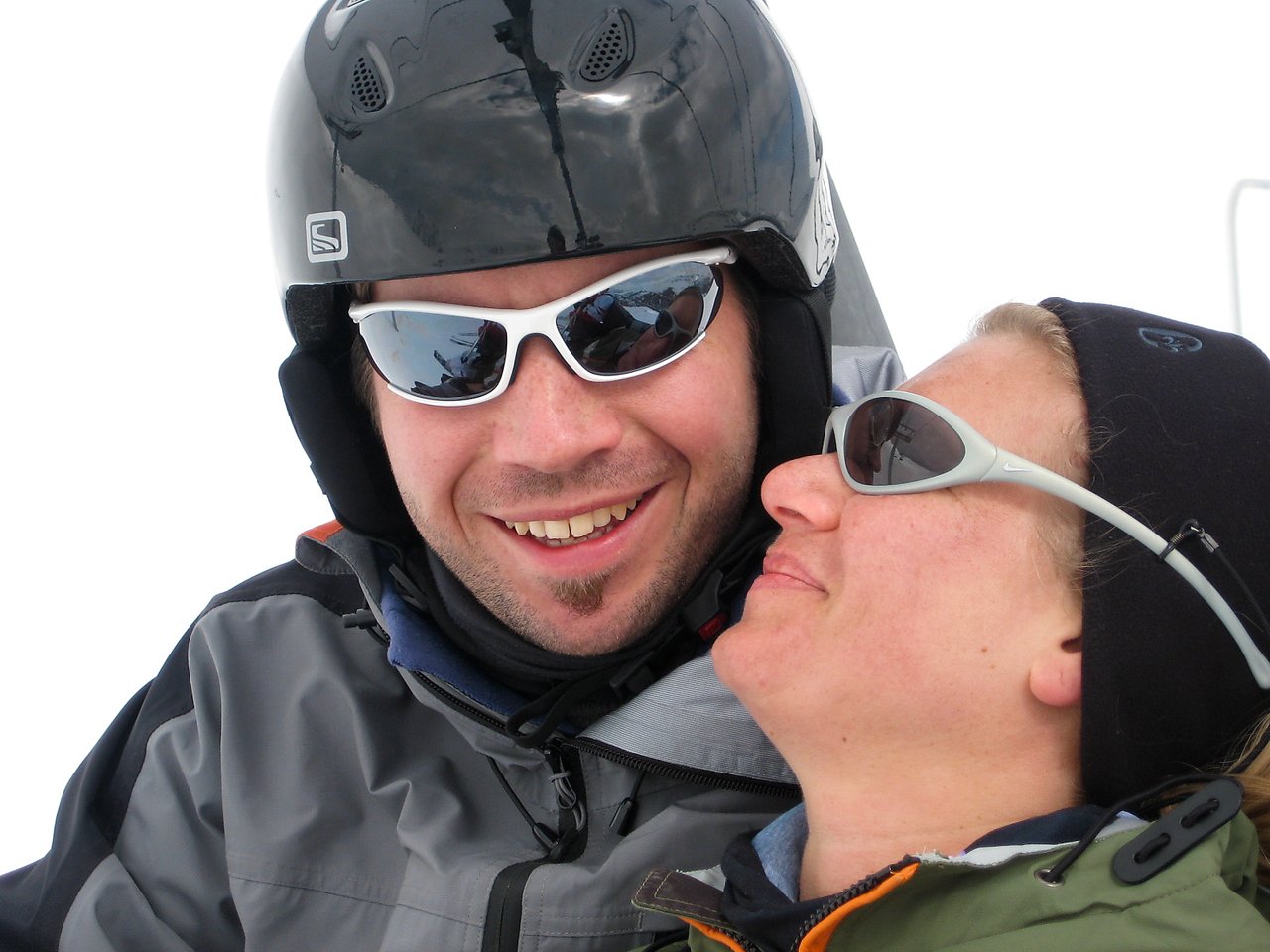 A man in a helmet and sunglasses smiles while a woman in sunglasses leans close to him.