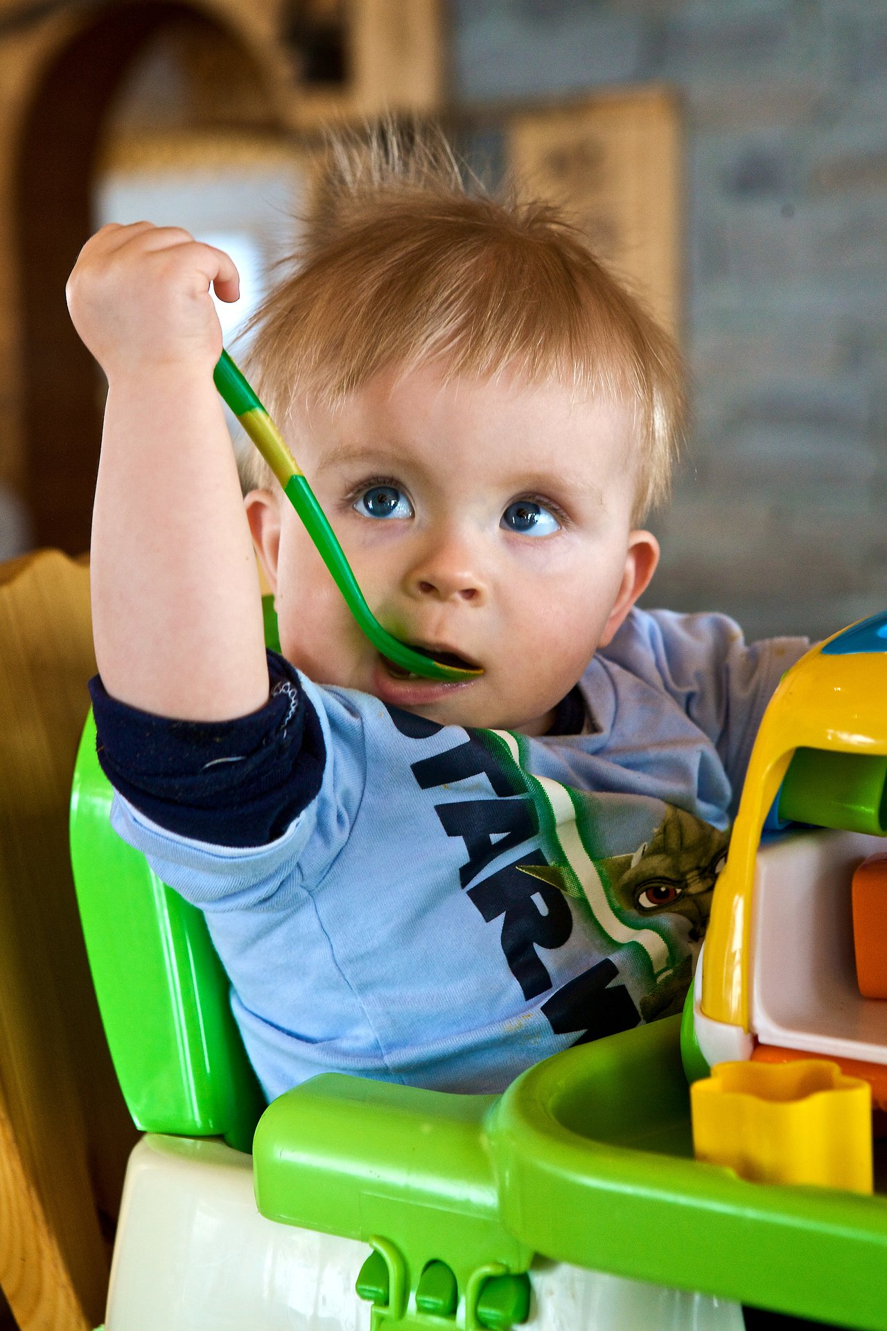 A baby in a high chair holds a green spoon and puts it in their mouth while looking up.