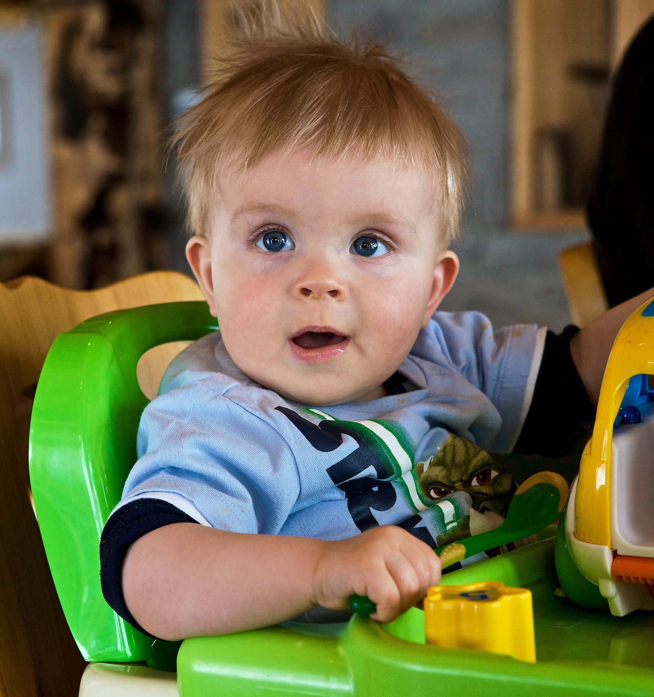 A baby with light hair and blue eyes sits in a green high chair, holding a toy and looking ahead.