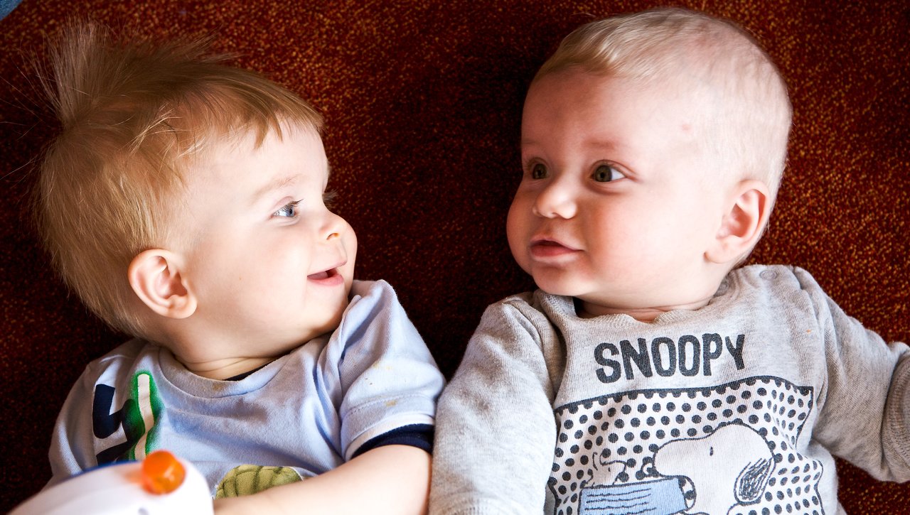 Two babies lie on a red carpet, facing each other.
