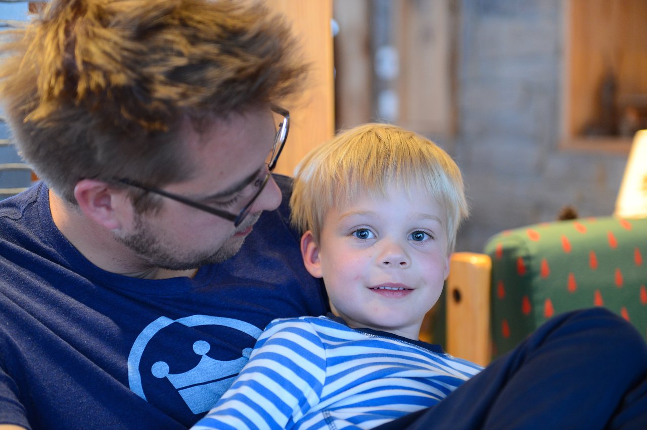 A father and his young son sit together, with the child smiling at the camera while the father looks at him.