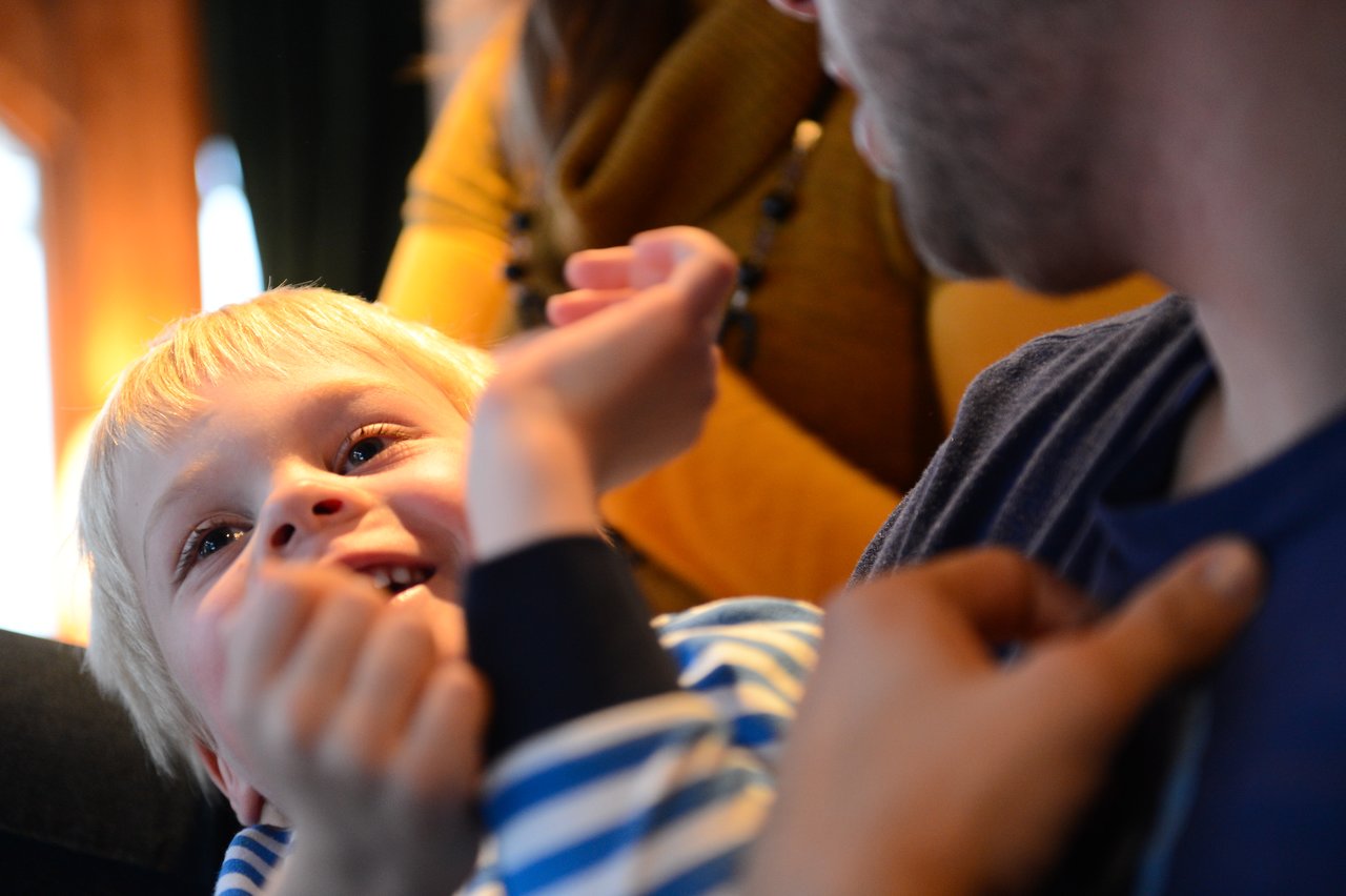 A young child smiles and looks up while playfully interacting with an adult sitting close by.
